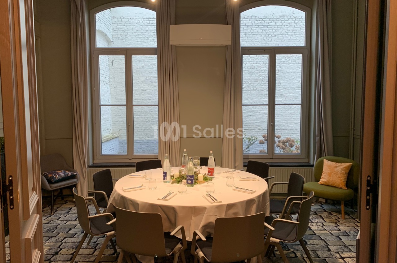 Salle lumineuse avec une table ronde dressée, entourée de chaises, devant de grandes fenêtres donnant sur un mur en briques…