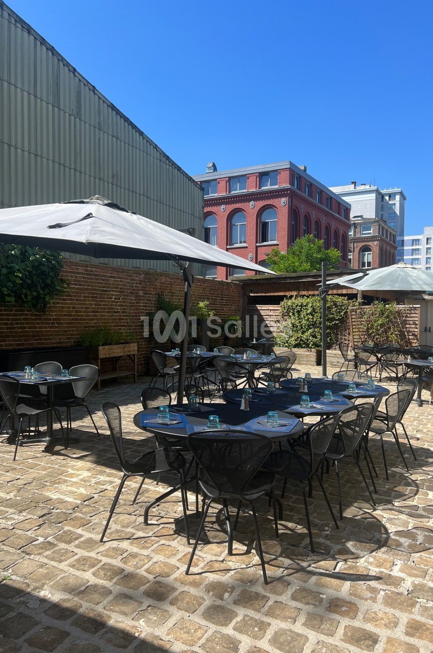 Terrasse extérieure avec tables et chaises en métal, parasols ouverts, entourée de murs en briques et bâtiments urbains.