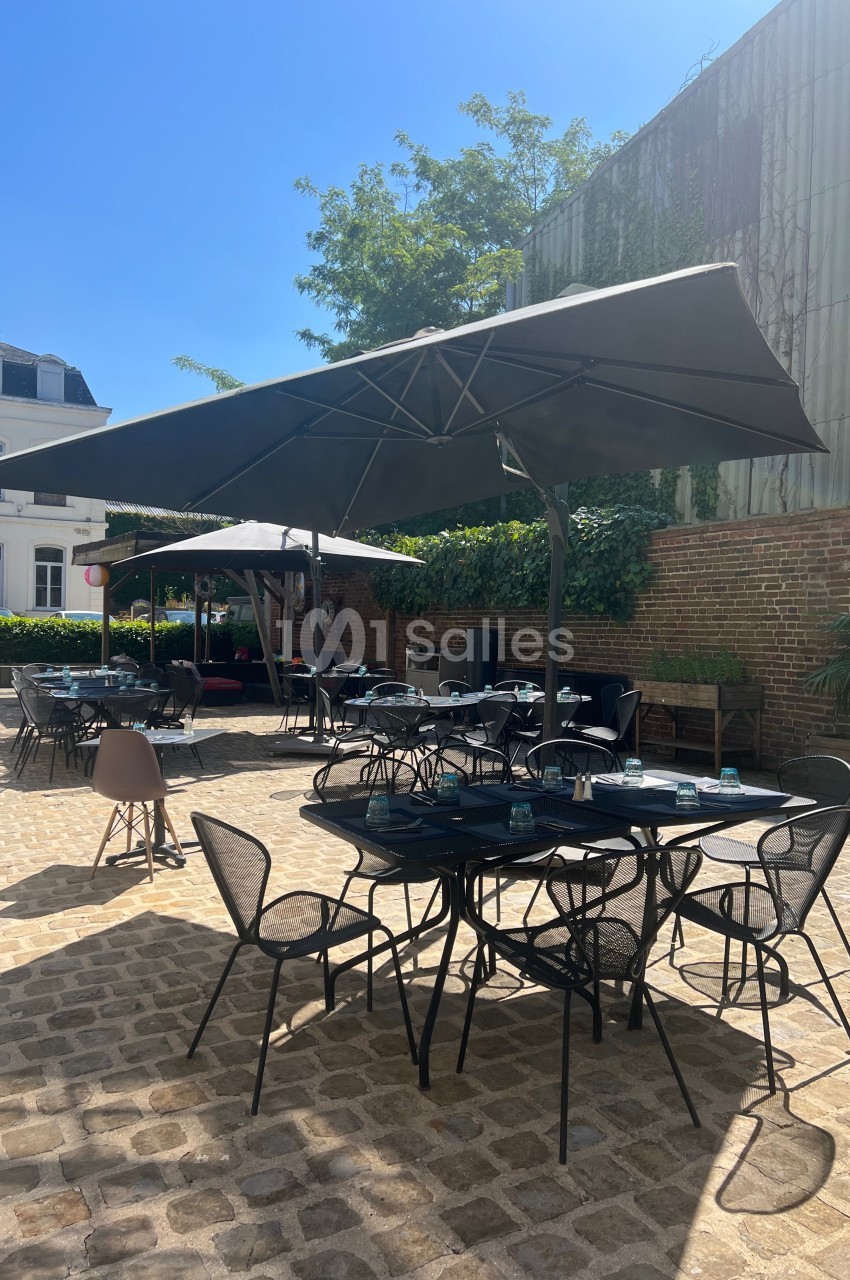 Terrasse extérieure ensoleillée avec tables et chaises noires, parasols ouverts et murs en briques en arrière-plan.