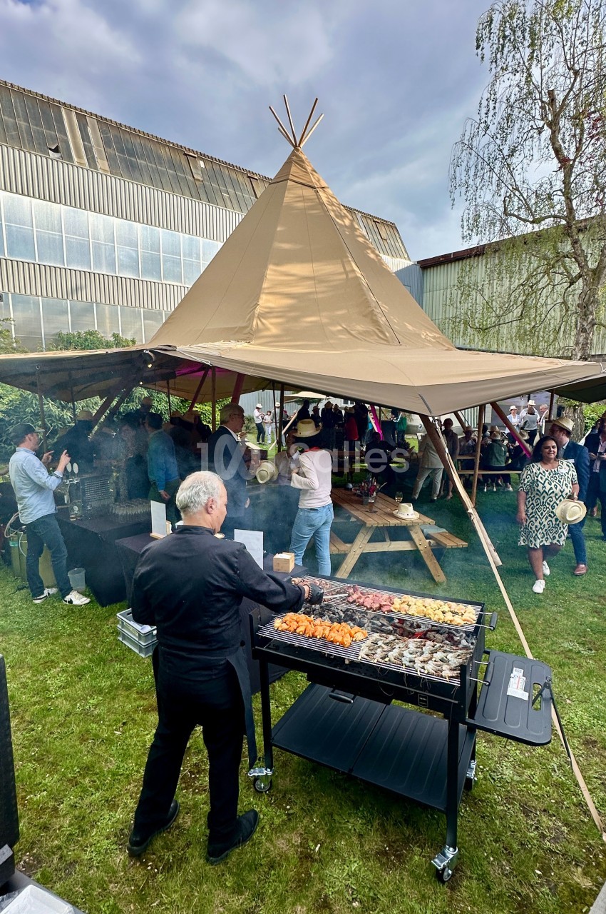 Un cuisinier prépare des brochettes sur un barbecue lors d'un événement en plein air sous une tente tipi.