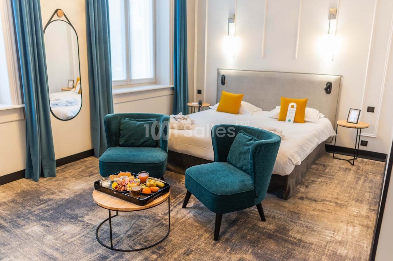 Chambre d'hôtel lumineuse avec lit double, fauteuils verts, table basse et plateau de petit-déjeuner.