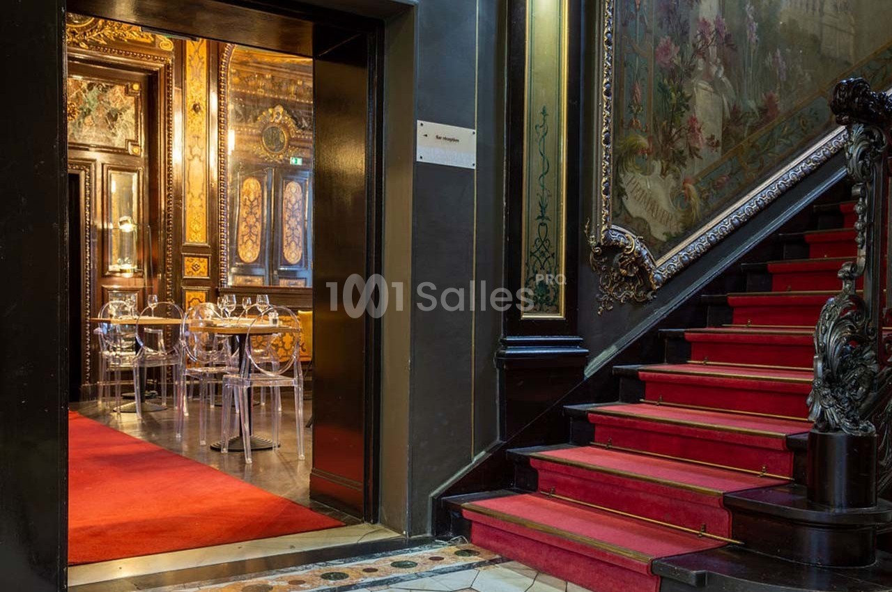 Escalier en tapis rouge menant à une salle ornée de décorations murales et de chaises transparentes.