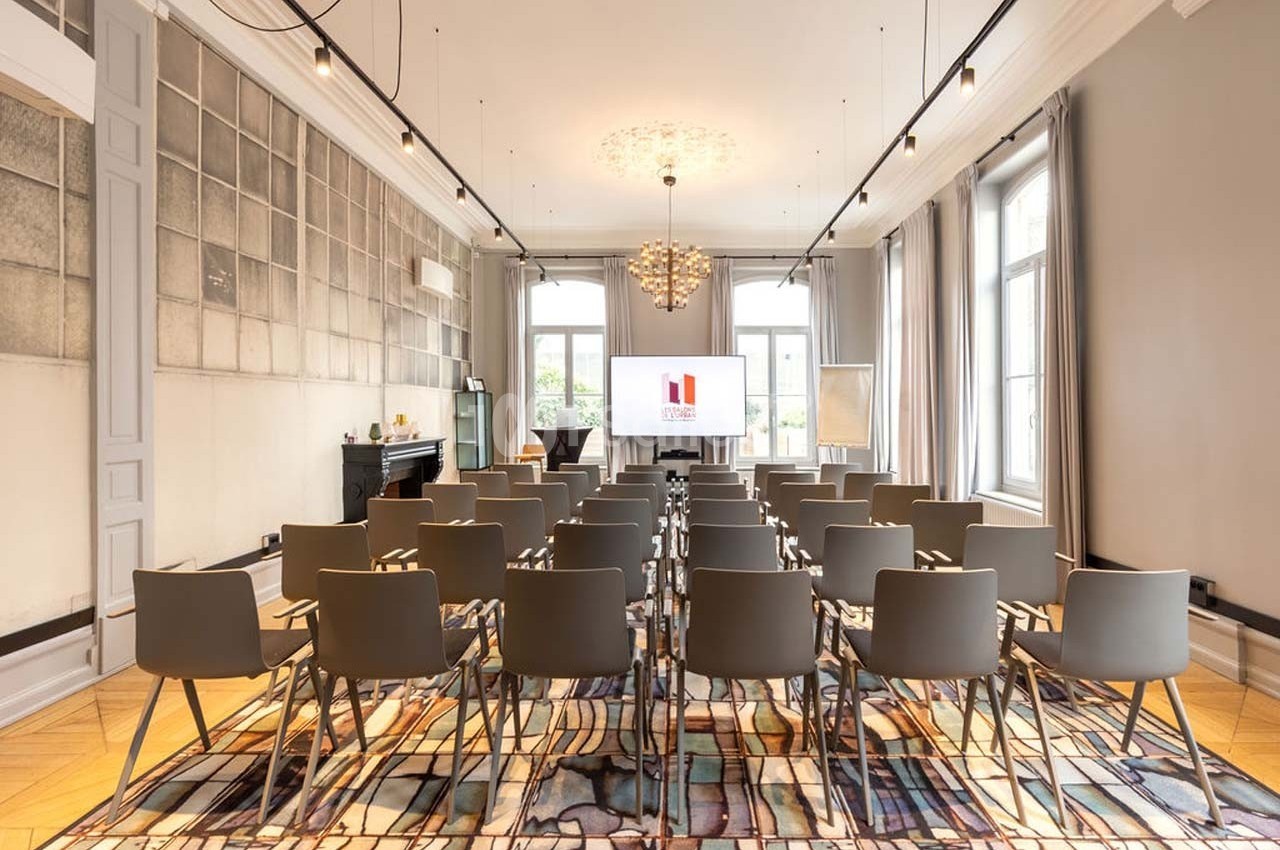 Salle de conférence lumineuse avec des rangées de chaises grises, un écran de projection et de grandes fenêtres.