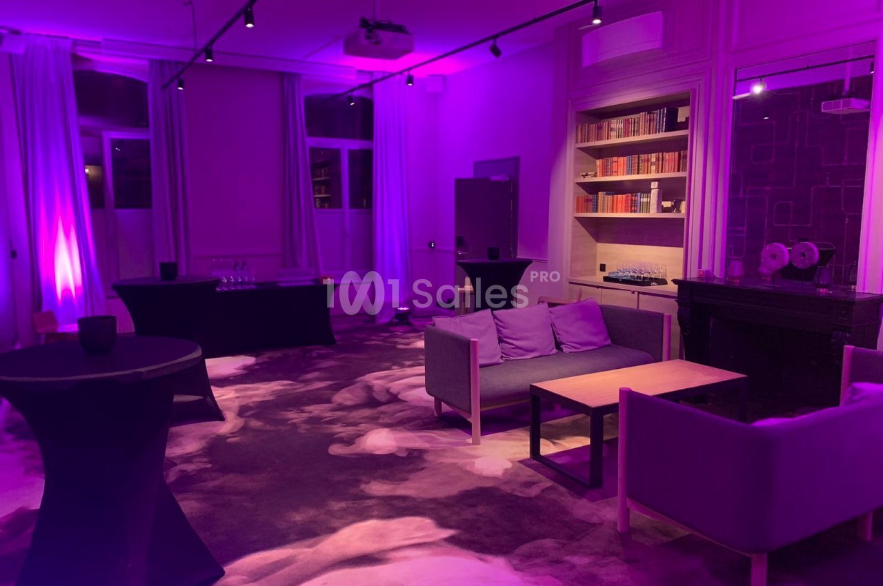 Salle de réception avec éclairage violet, canapés, tables basses et étagères remplies de livres en arrière-plan.