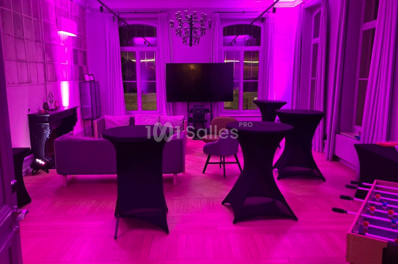 Salle aménagée avec des tables hautes, un canapé, un écran et un éclairage rose dans un cadre intérieur élégant.