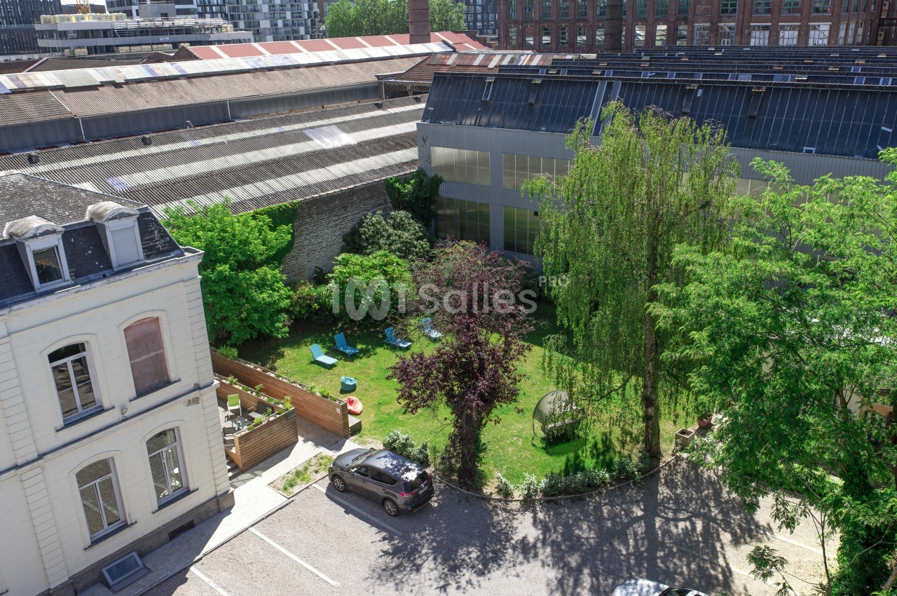 Vue d'un jardin verdoyant avec des arbres, des transats et des bâtiments industriels en arrière-plan.