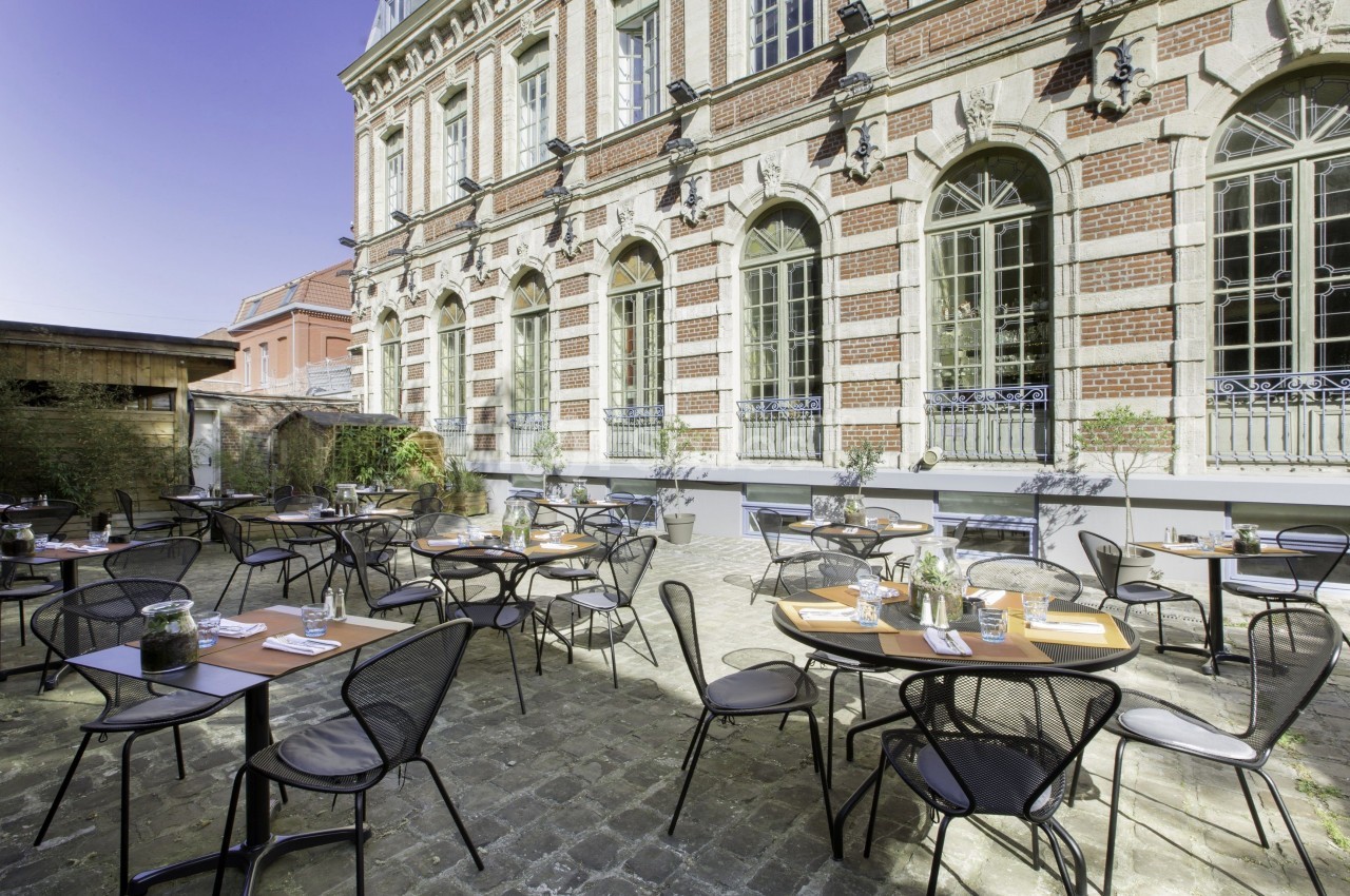 Terrasse extérieure avec tables et chaises en métal, située devant un bâtiment en briques avec de grandes fenêtres.