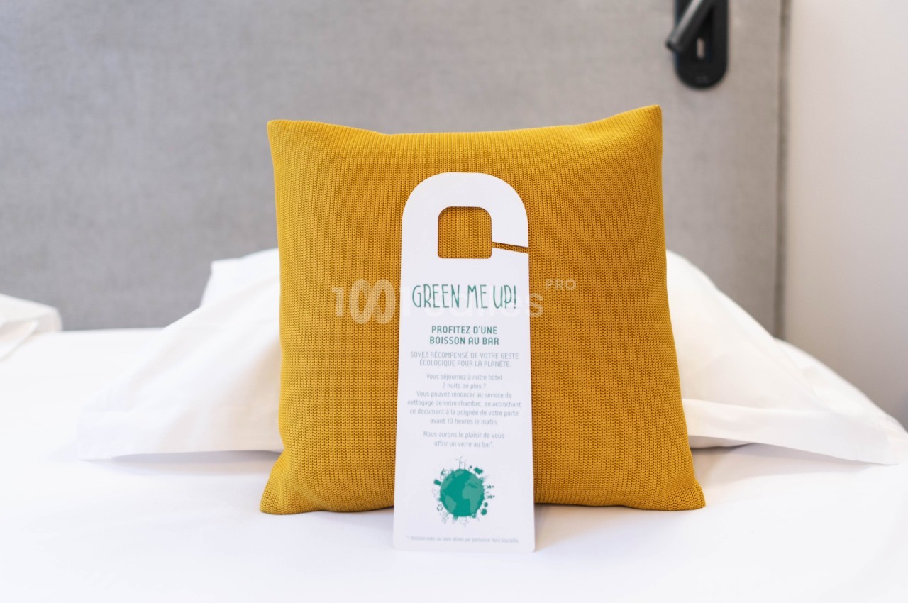 Coussin jaune sur un lit blanc avec une pancarte ’Green Me Up!’ suspendue, promouvant une initiative écologique.