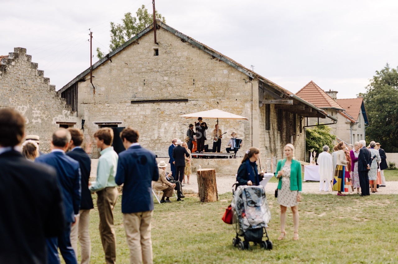 Groupe de personnes rassemblées en extérieur devant un bâtiment en pierre, avec des musiciens jouant sur une petite scène.