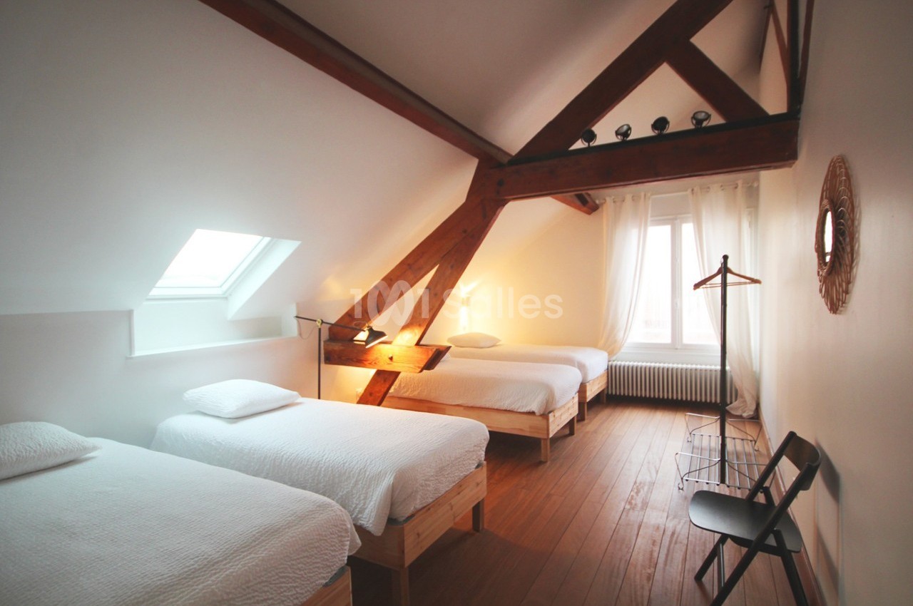 Chambre mansardée avec trois lits simples, poutres apparentes, lumière naturelle et mobilier minimaliste.