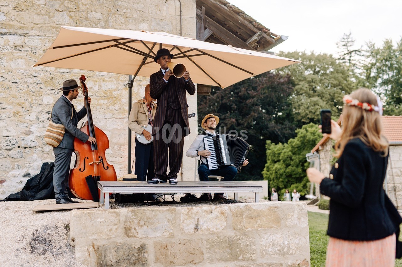 Un groupe de musiciens joue en plein air sous un parasol, avec une femme prenant une photo au premier plan.
