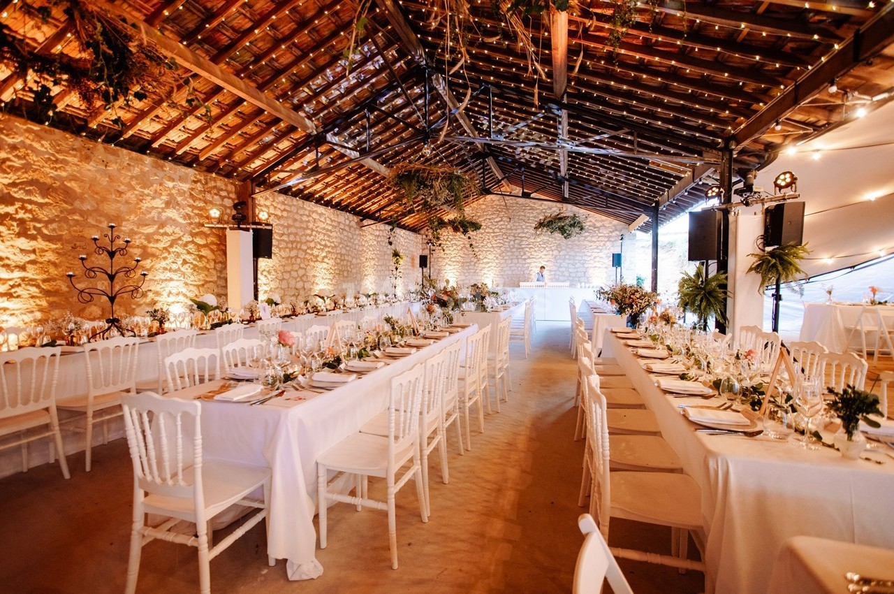 Salle de réception décorée avec des tables alignées, nappes blanches, chaises et guirlandes lumineuses suspendues.