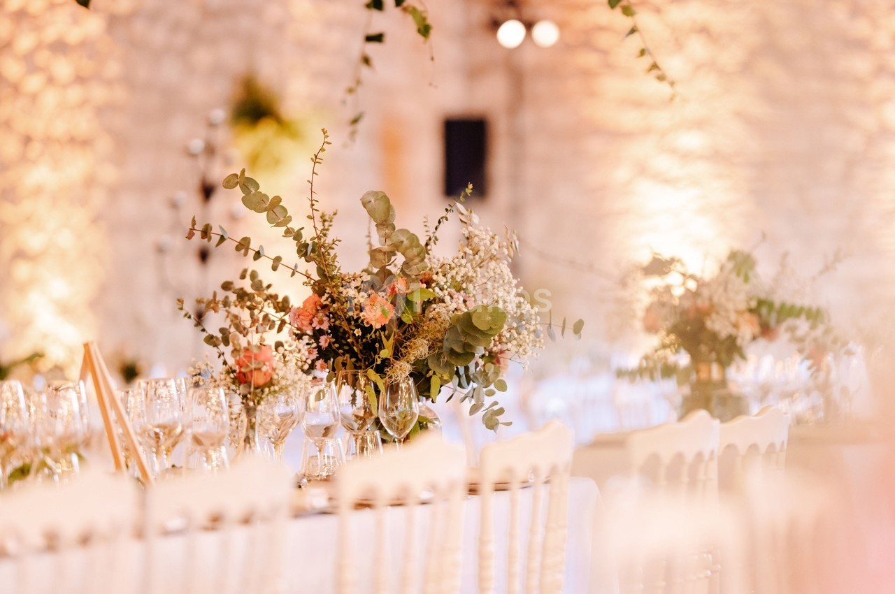 Décoration florale avec des bouquets de fleurs et feuillages sur des tables dressées dans une salle en pierre éclairée.