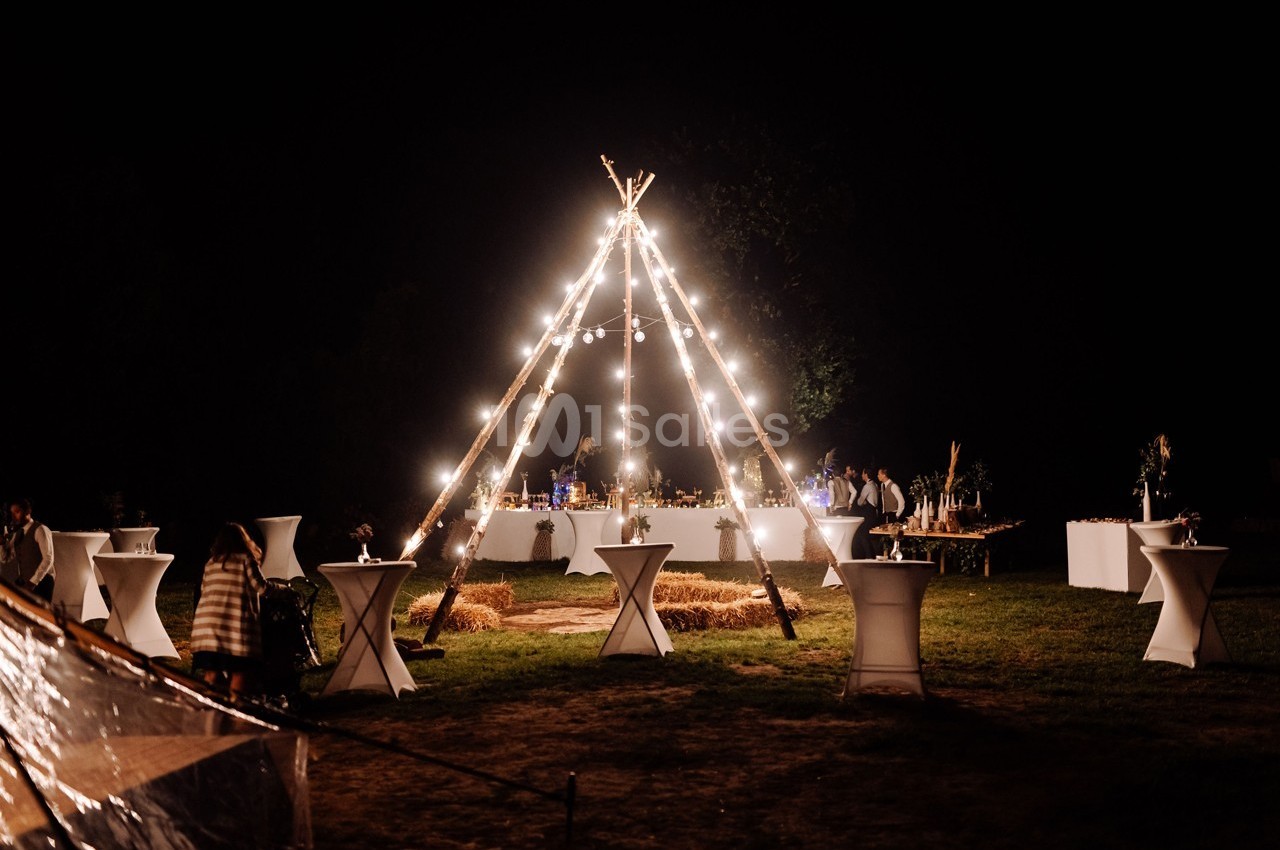 Tente tipi éclairée par des guirlandes lumineuses, entourée de tables hautes dans un décor nocturne en extérieur.