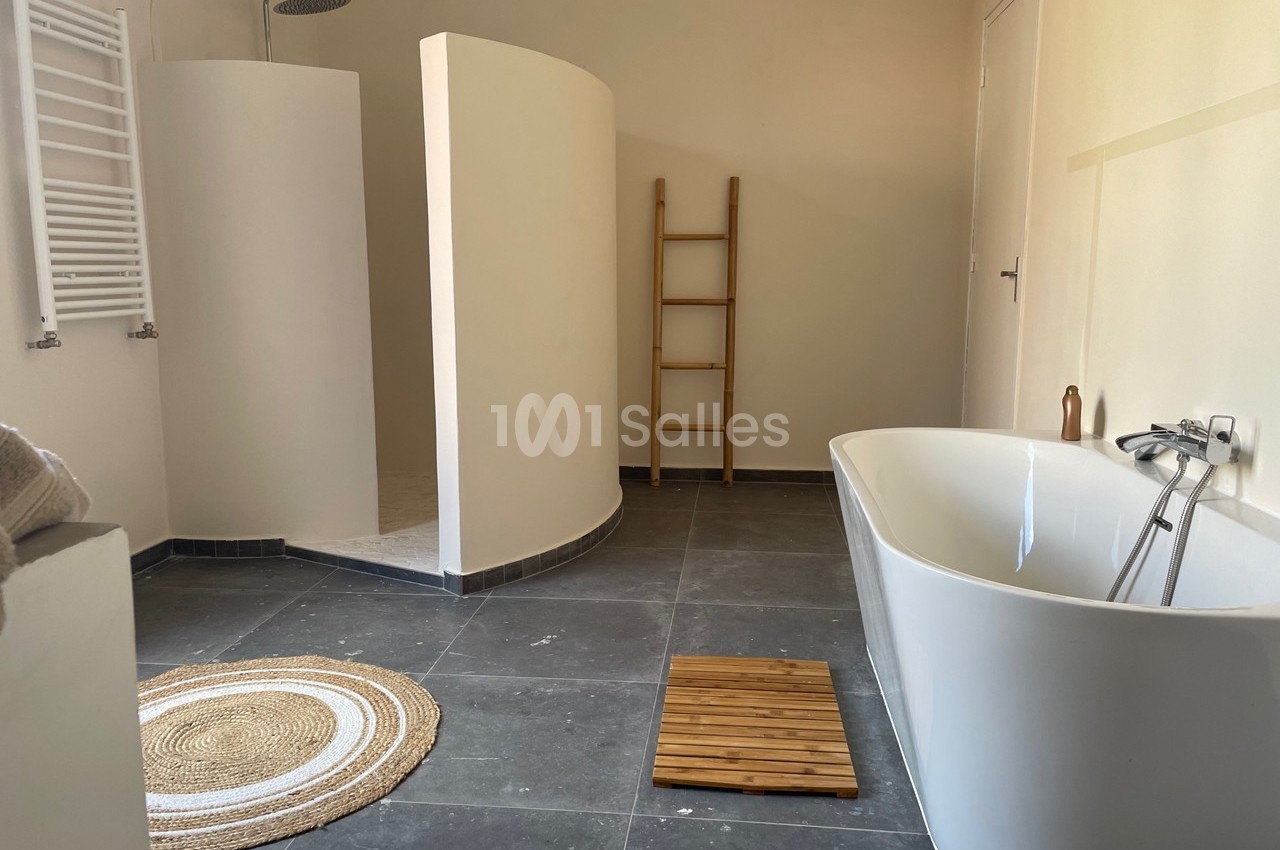 Salle de bain moderne avec douche ouverte, baignoire blanche, tapis rond en jute et échelle en bois décorative.