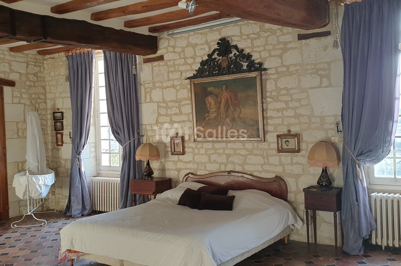 Chambre avec murs en pierre, poutres apparentes, lit double, rideaux violets et mobilier ancien.