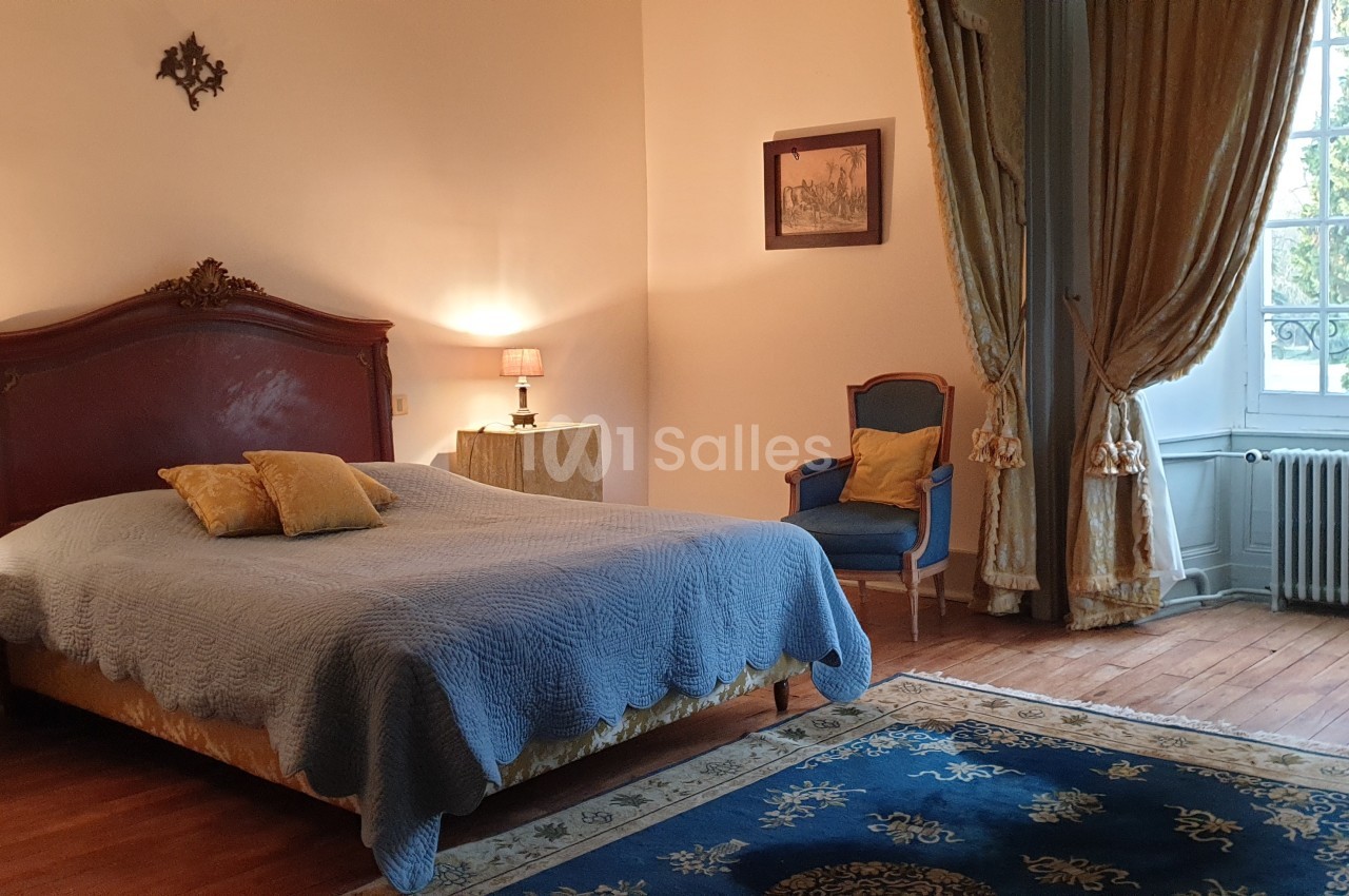 Chambre lumineuse avec lit double, fauteuil, tapis bleu, rideaux dorés et grande fenêtre donnant sur un jardin.