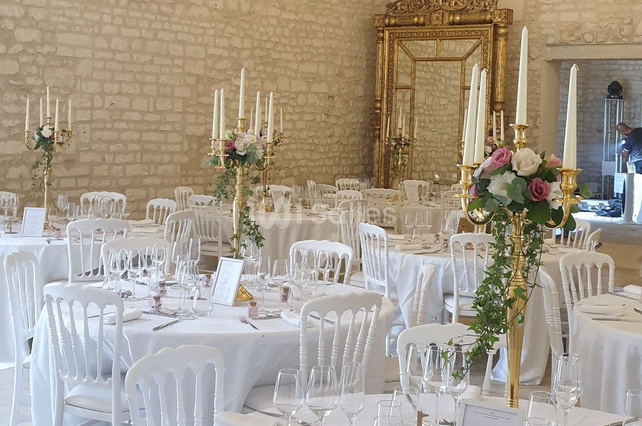Salle de réception décorée avec des tables rondes, nappes blanches, chandeliers dorés et compositions florales.