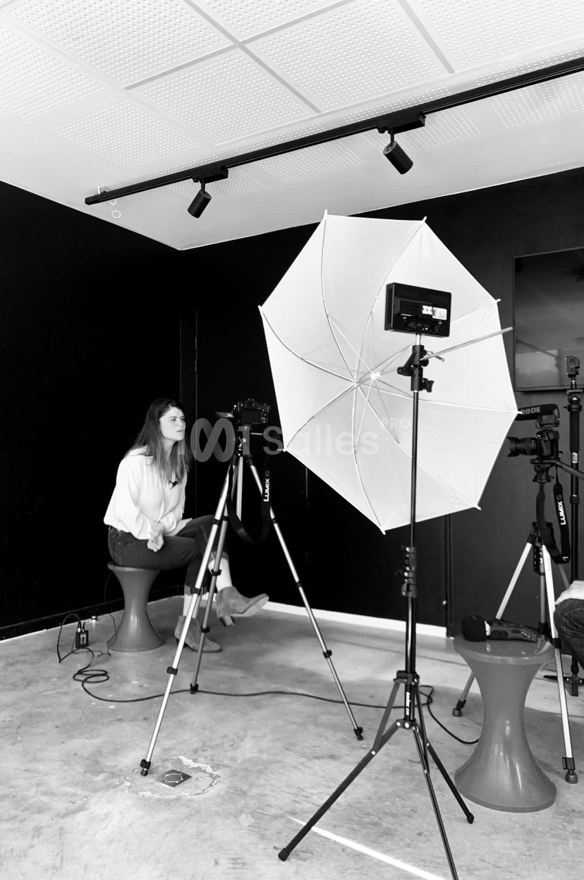 Une femme assise devant un appareil photo sur trépied, éclairée par un parapluie lumineux dans un studio sombre.
