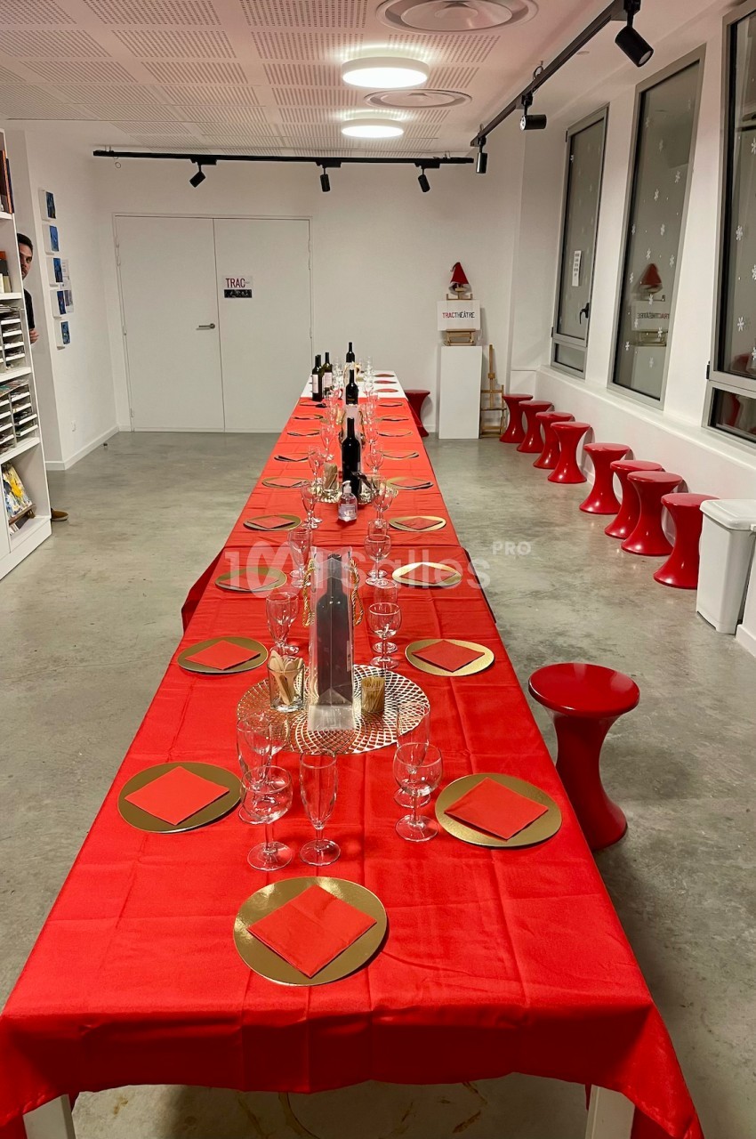 Grande table rectangulaire décorée pour un repas festif, avec nappes rouges, verres et assiettes dorées dans une salle…