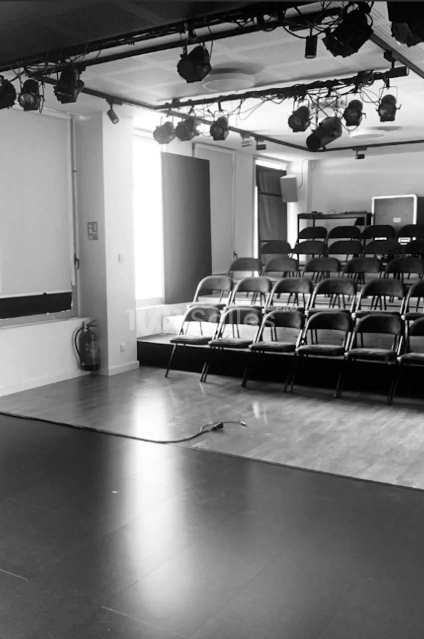 Salle de spectacle vide avec des rangées de chaises noires face à une scène, éclairage suspendu au plafond.
