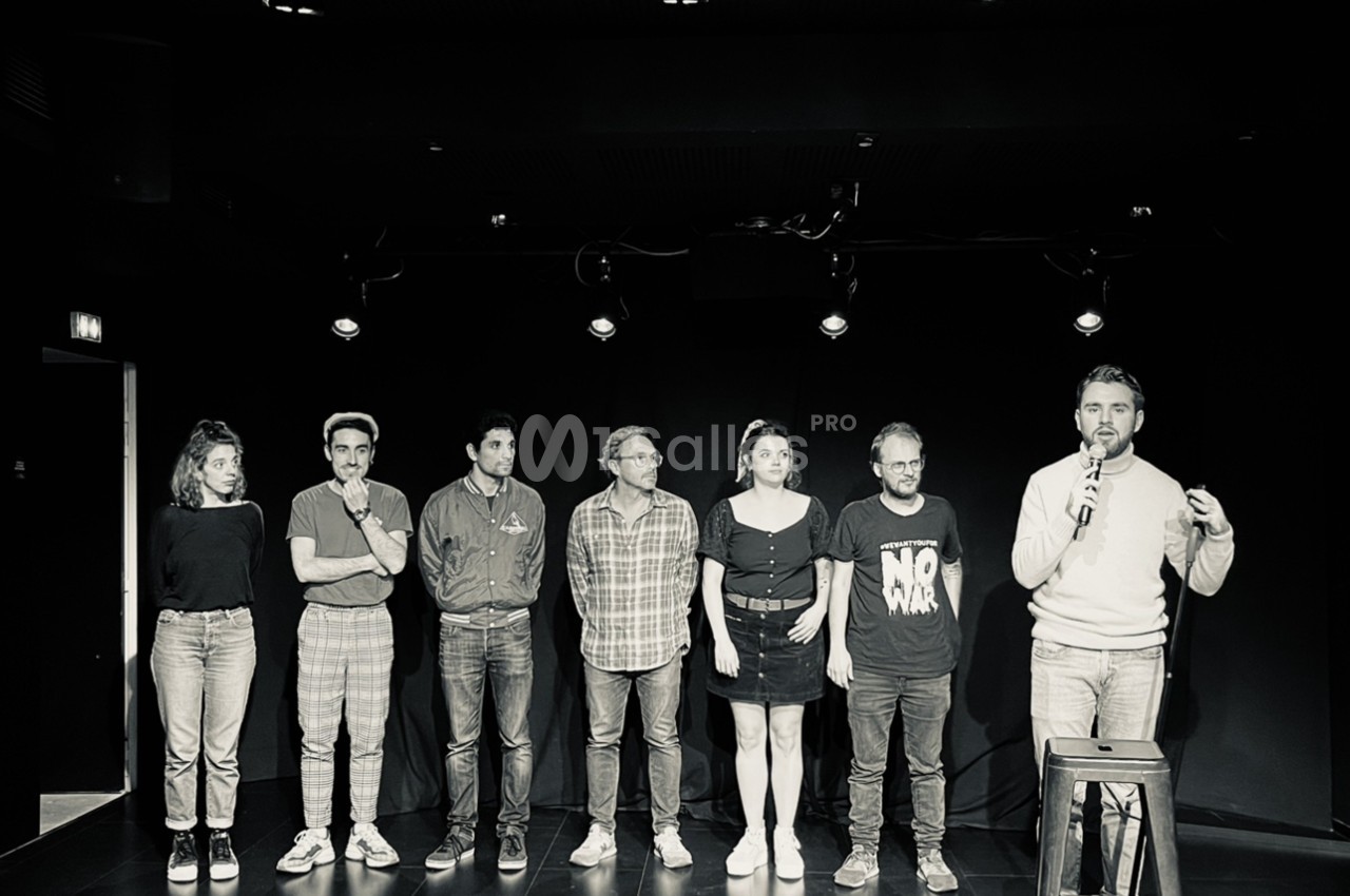 Un groupe de sept personnes debout sur une scène sombre, un homme tenant un micro à droite.