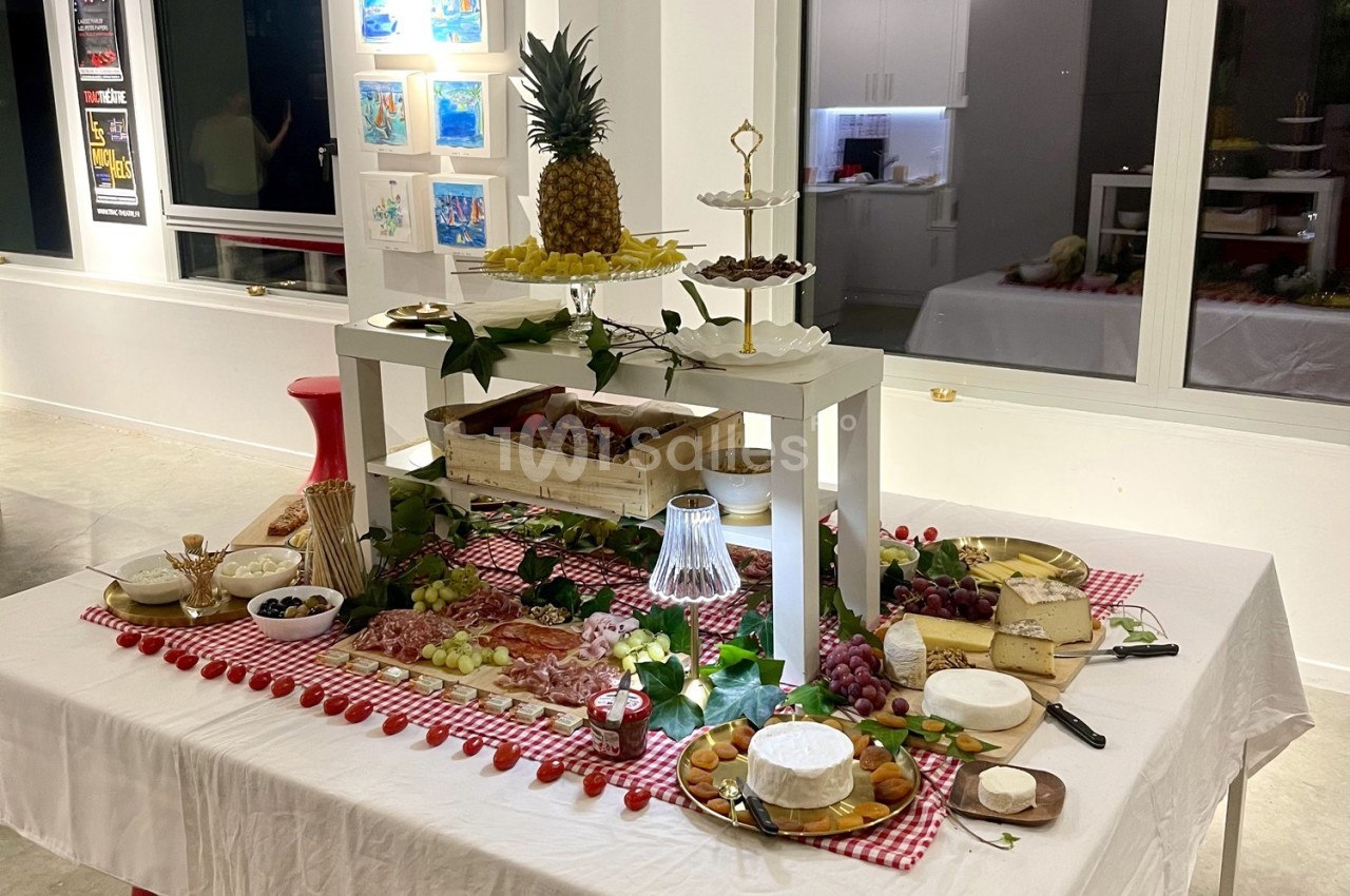 Table dressée avec des fromages, charcuteries, fruits et desserts, décorée de nappes à carreaux rouges et éléments végétaux.