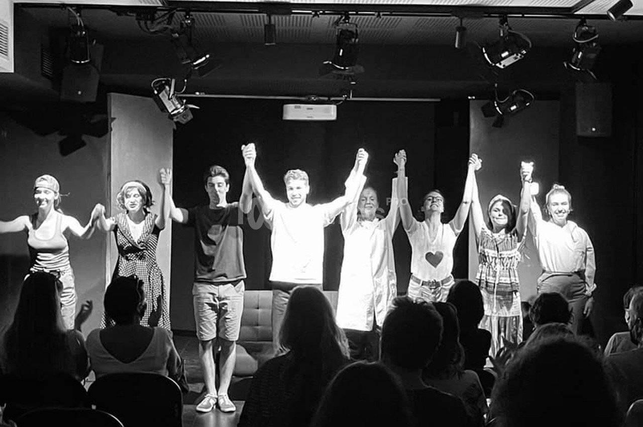 Des comédiens saluent le public sur une scène de théâtre, éclairés par des projecteurs, devant une audience attentive.