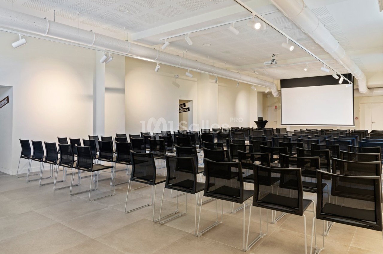 Salle de conférence lumineuse avec des rangées de chaises noires et un écran de projection blanc à l'avant.