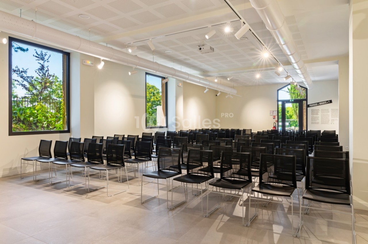 Salle lumineuse avec des rangées de chaises noires alignées, grandes fenêtres donnant sur un espace verdoyant.
