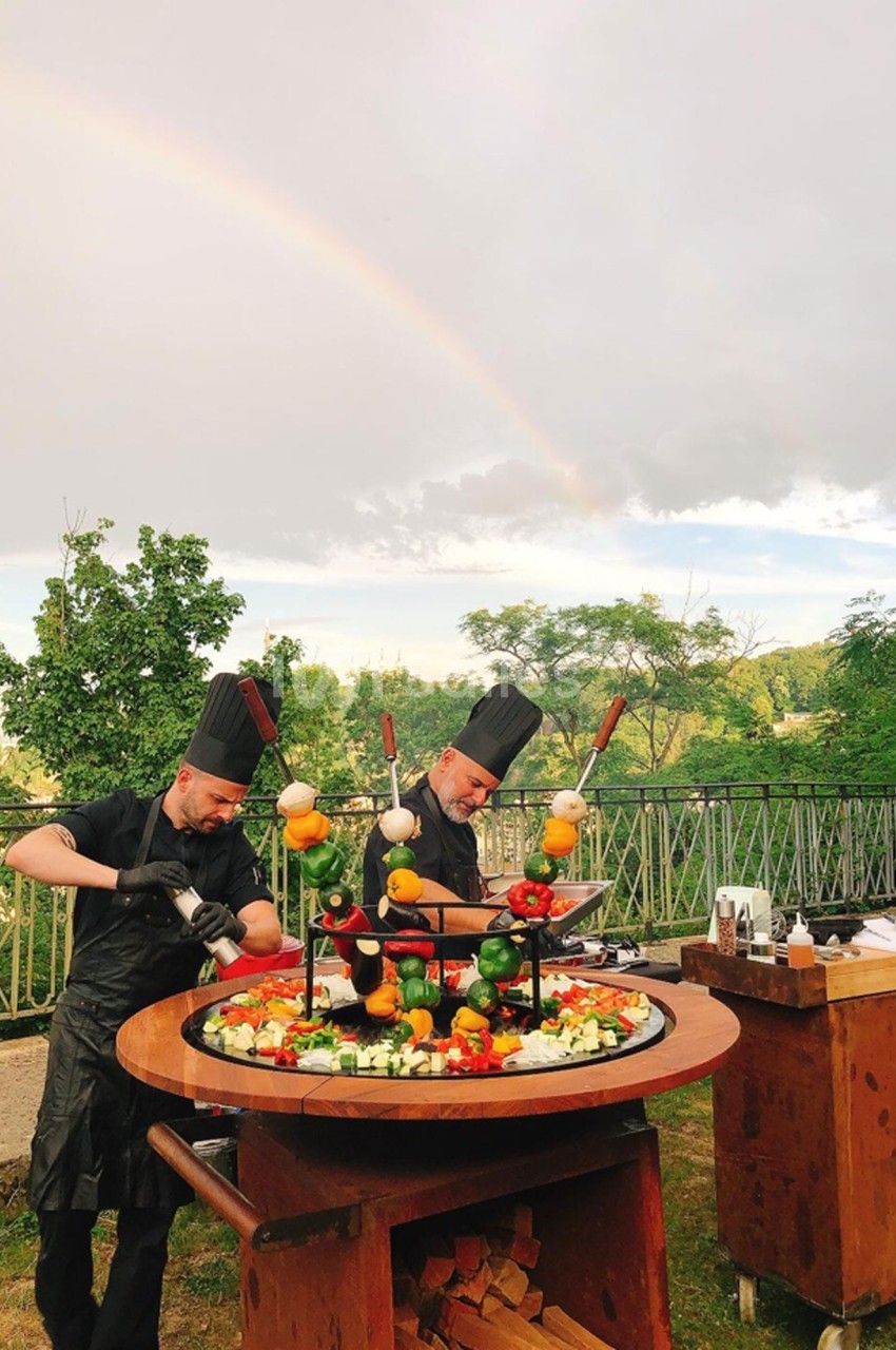 Deux chefs cuisinent des brochettes de légumes et de viande sur un barbecue circulaire en plein air sous un arc-en-ciel.