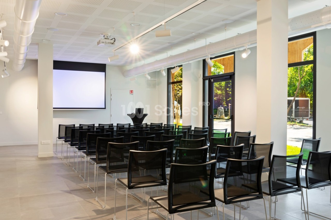 Salle de conférence lumineuse avec des chaises noires alignées, un écran de projection et de grandes fenêtres donnant sur l…