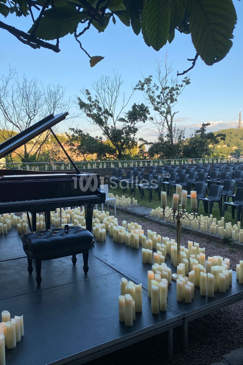 Scène extérieure avec un piano à queue entouré de bougies, des rangées de chaises vides en arrière-plan.