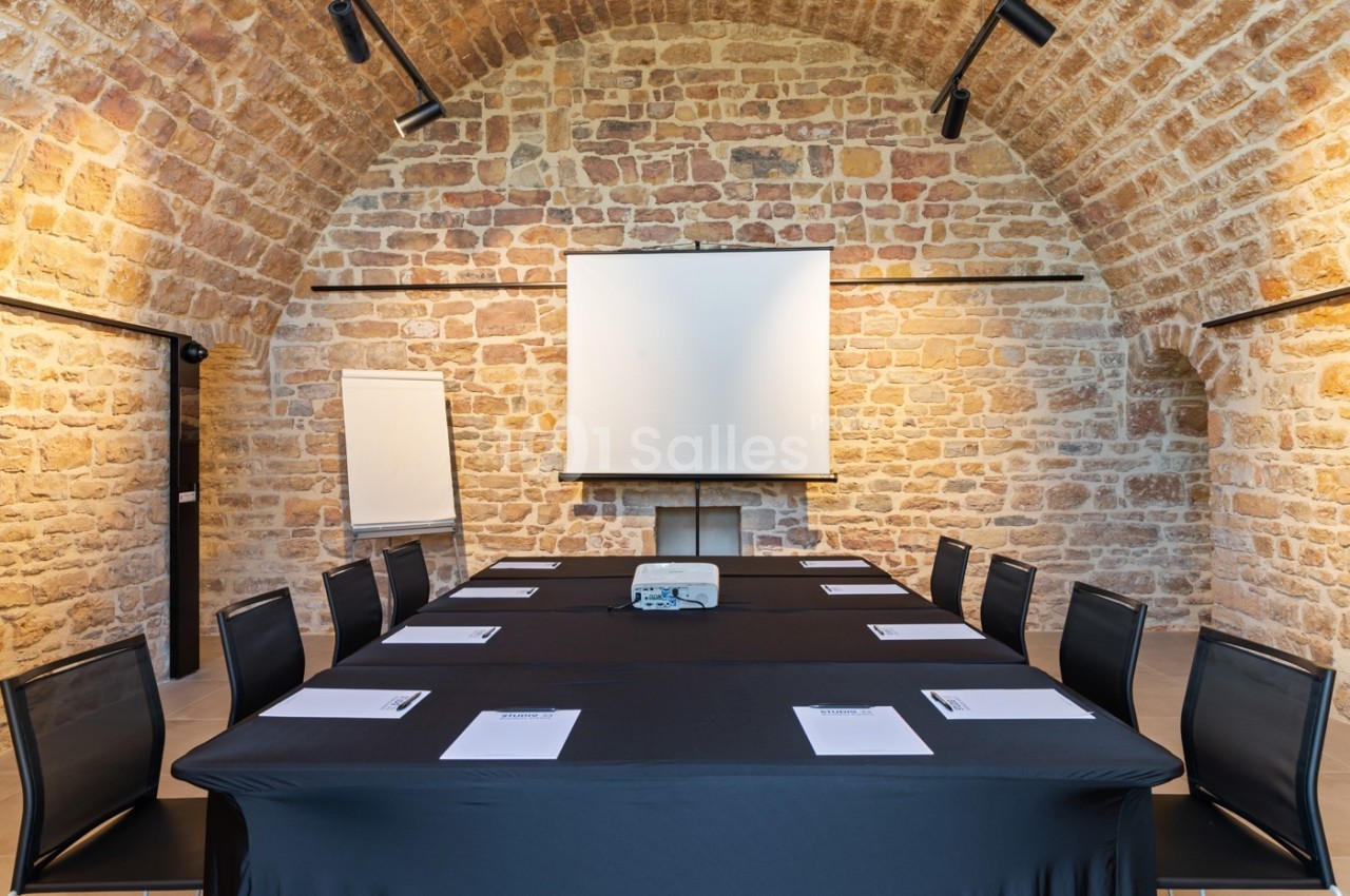 Salle de réunion avec table rectangulaire, chaises noires, projecteur, écran blanc et murs en pierre apparente.
