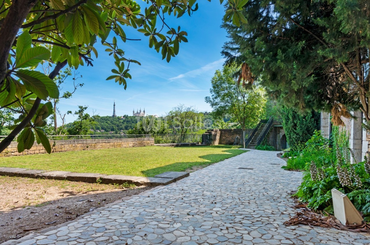 Allée pavée bordée de verdure menant à un espace ouvert avec vue sur une colline et des bâtiments au loin.