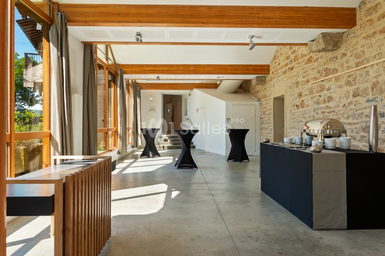 Salle lumineuse avec murs en pierre, grandes baies vitrées, tables hautes noires et buffet dressé sur la droite.