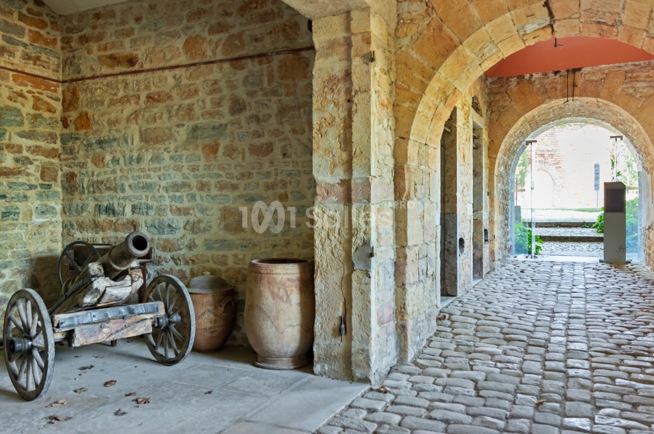 Un vieux canon sur roues et des jarres en terre cuite sont disposés sous une arcade en pierre dans une cour pavée.