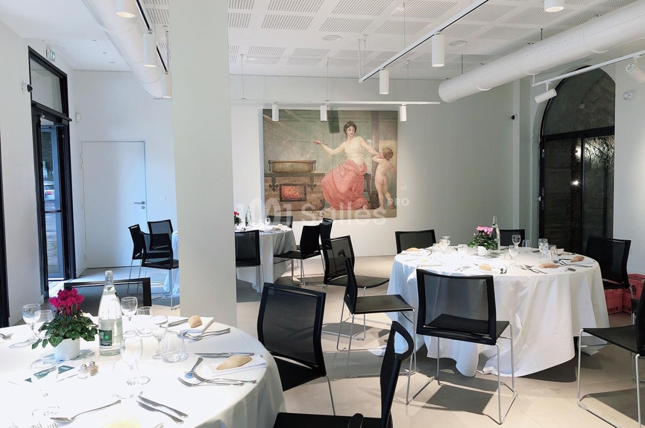 Salle lumineuse avec tables rondes dressées pour un repas, décorée d'une grande peinture murale et de fleurs.