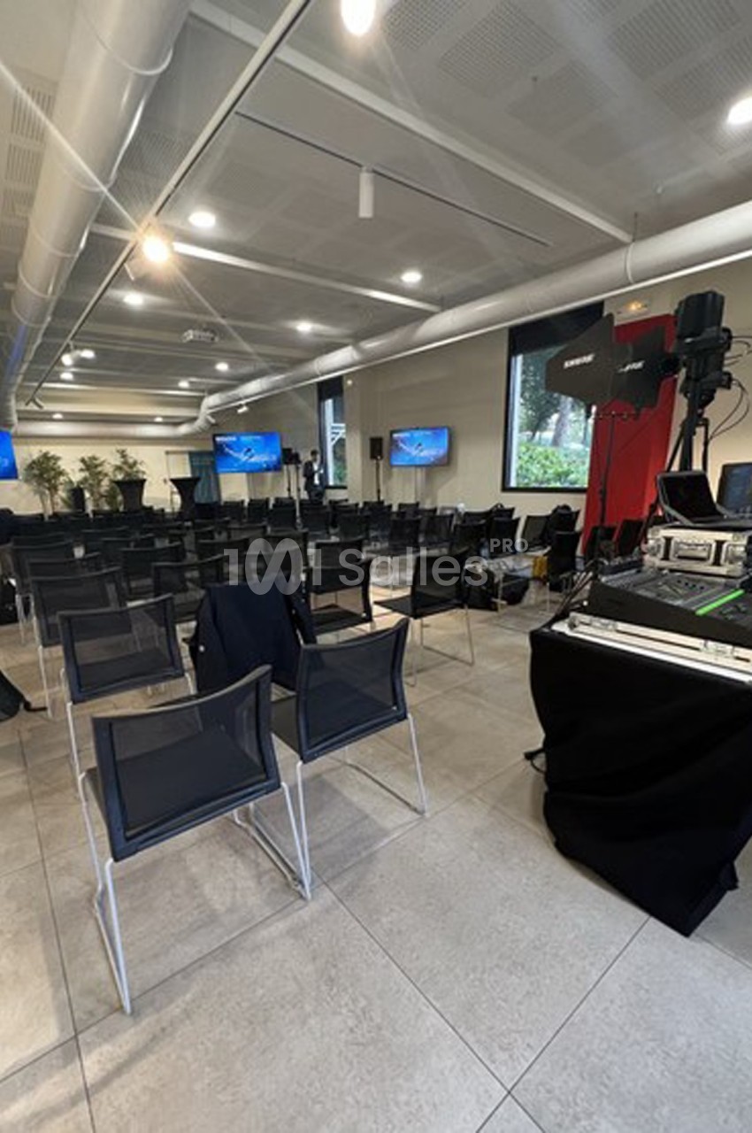 Salle de conférence vide avec des chaises alignées, des écrans allumés et du matériel audiovisuel installé.