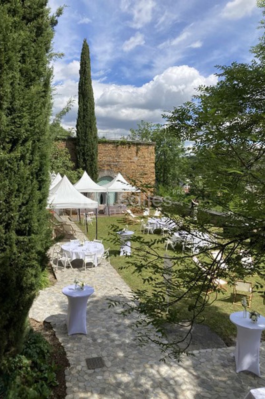 Jardin aménagé avec des tables et des tentes blanches, entouré de verdure et d'un mur en pierre sous un ciel partiellement…