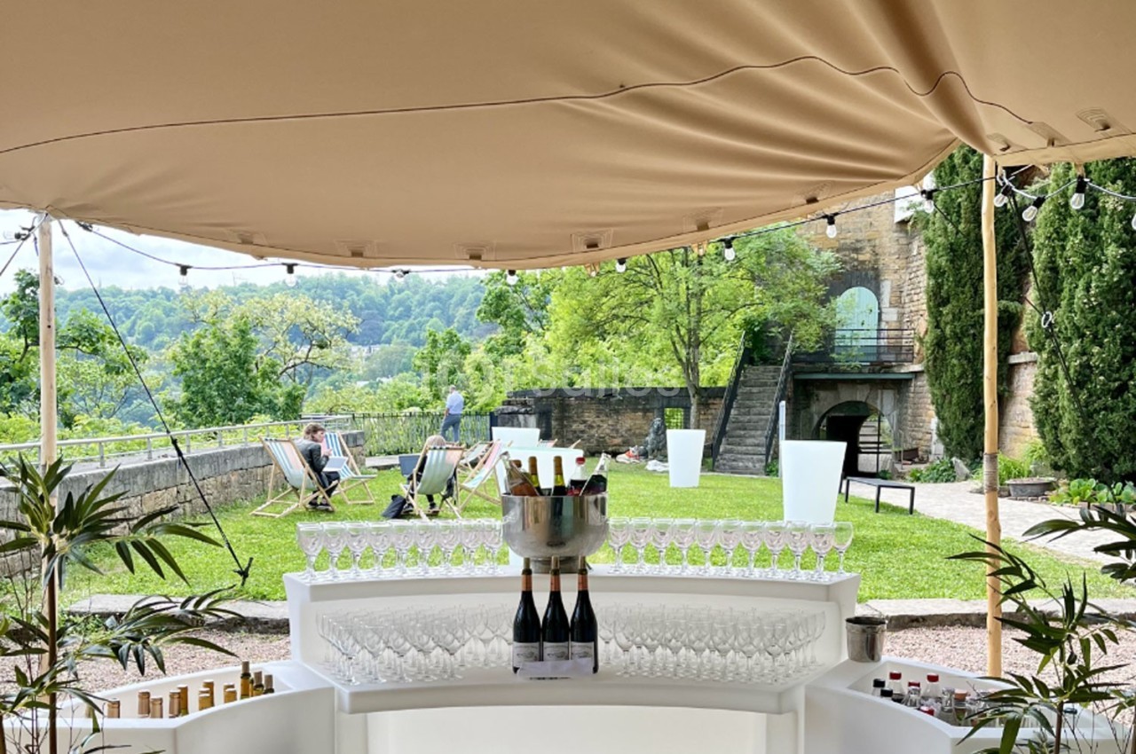 Bar extérieur sous une tente beige avec des verres et des bouteilles, vue sur un jardin verdoyant en arrière-plan.