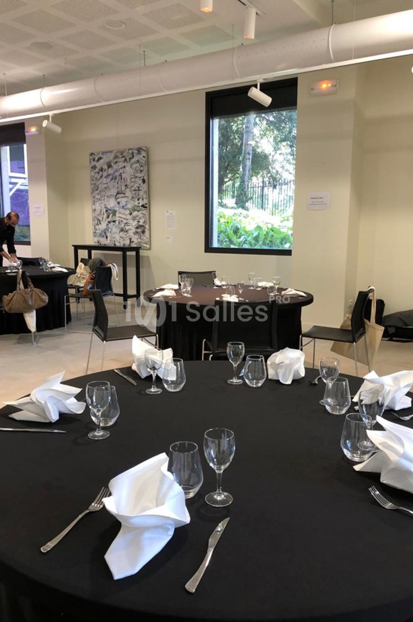 Tables rondes dressées avec nappes noires, serviettes pliées et verres dans une salle lumineuse avec grandes fenêtres.
