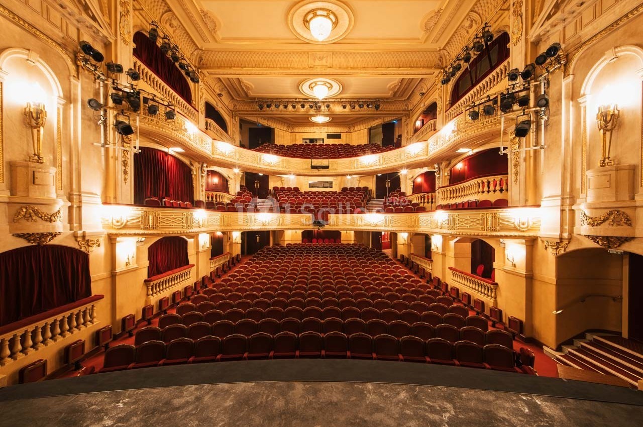 Vue intérieure d'un théâtre avec des rangées de sièges rouges, des balcons ornés et un éclairage chaleureux.