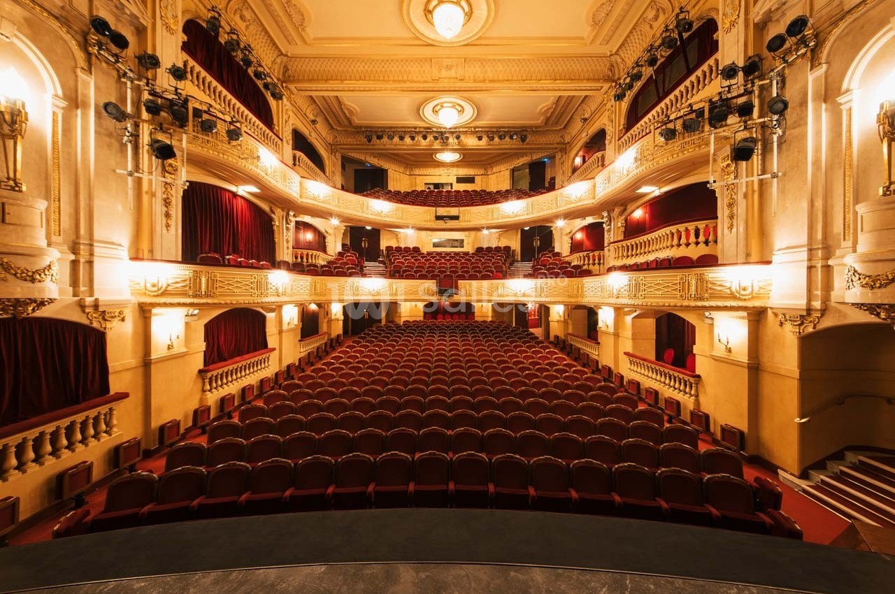 Salle de théâtre vide avec sièges rouges, balcons ornés et éclairage chaleureux, vue depuis la scène.