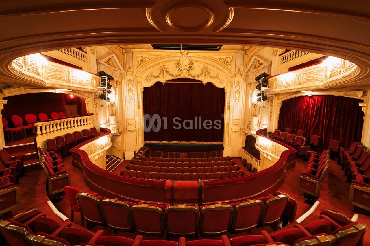Intérieur d'un théâtre avec des sièges rouges, une scène centrale avec rideau fermé et une architecture ornée.