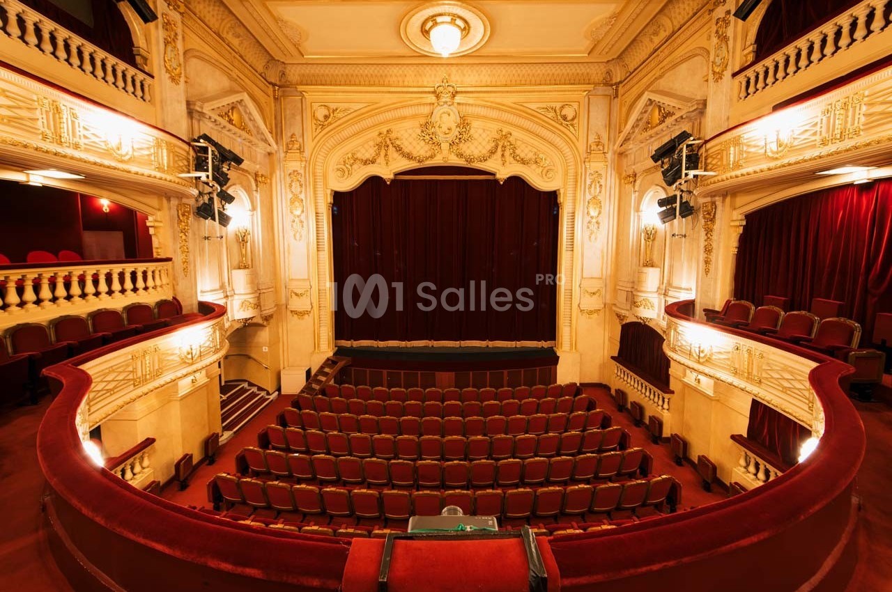 Intérieur d'un théâtre avec une scène fermée par un rideau rouge, entourée de balcons et de sièges vides.