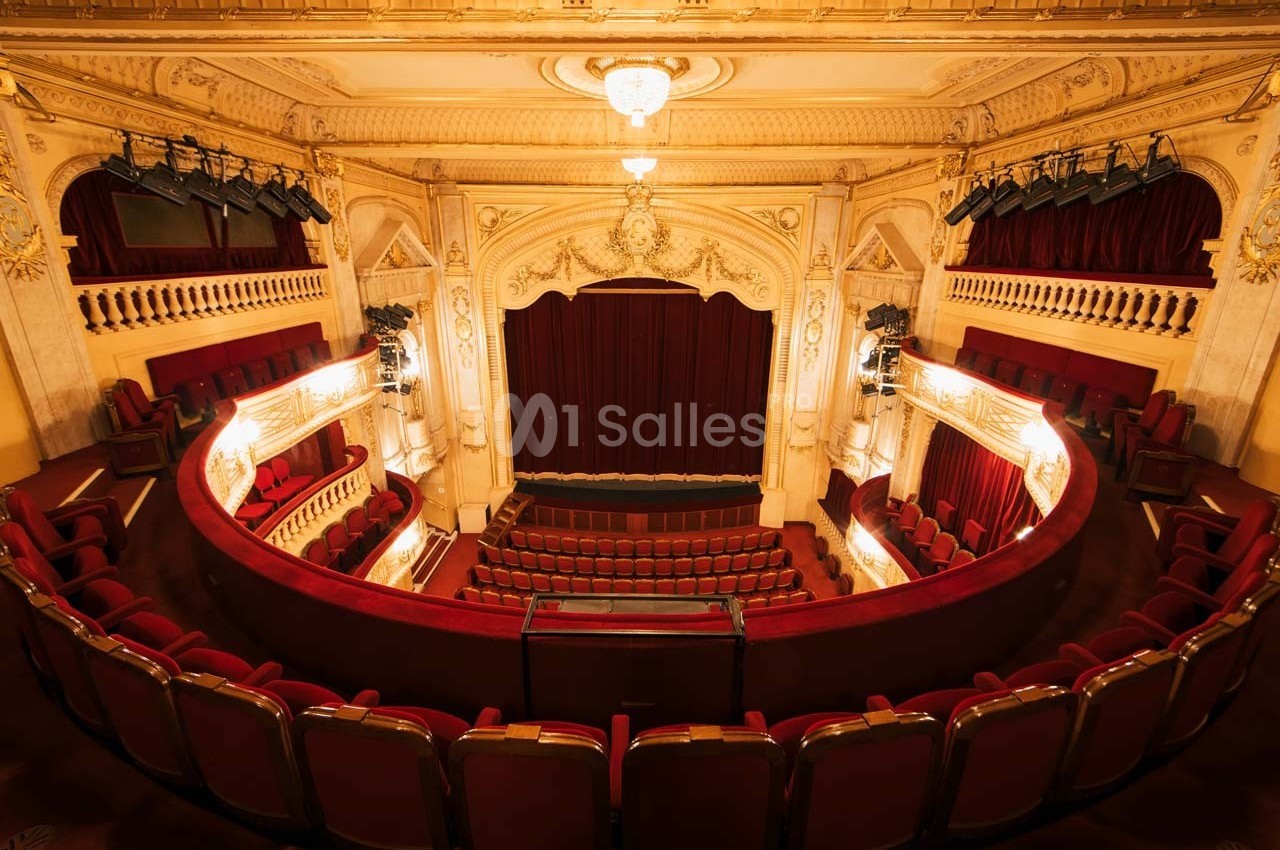 Intérieur d'un théâtre avec des sièges rouges, une scène encadrée de rideaux et un balcon décoré de moulures.