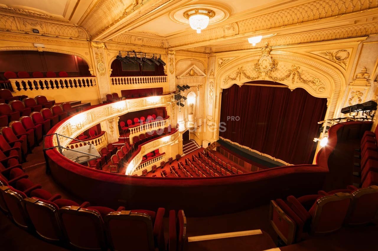 Salle de théâtre avec balcons ornés, sièges rouges et rideau de scène fermé dans un décor intérieur élégant.