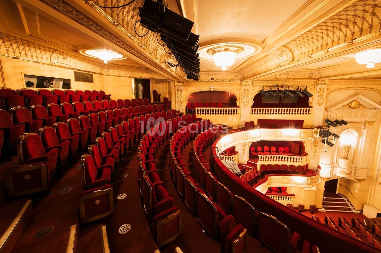 Salle de théâtre avec des rangées de sièges rouges, balcons décorés et éclairage chaleureux.