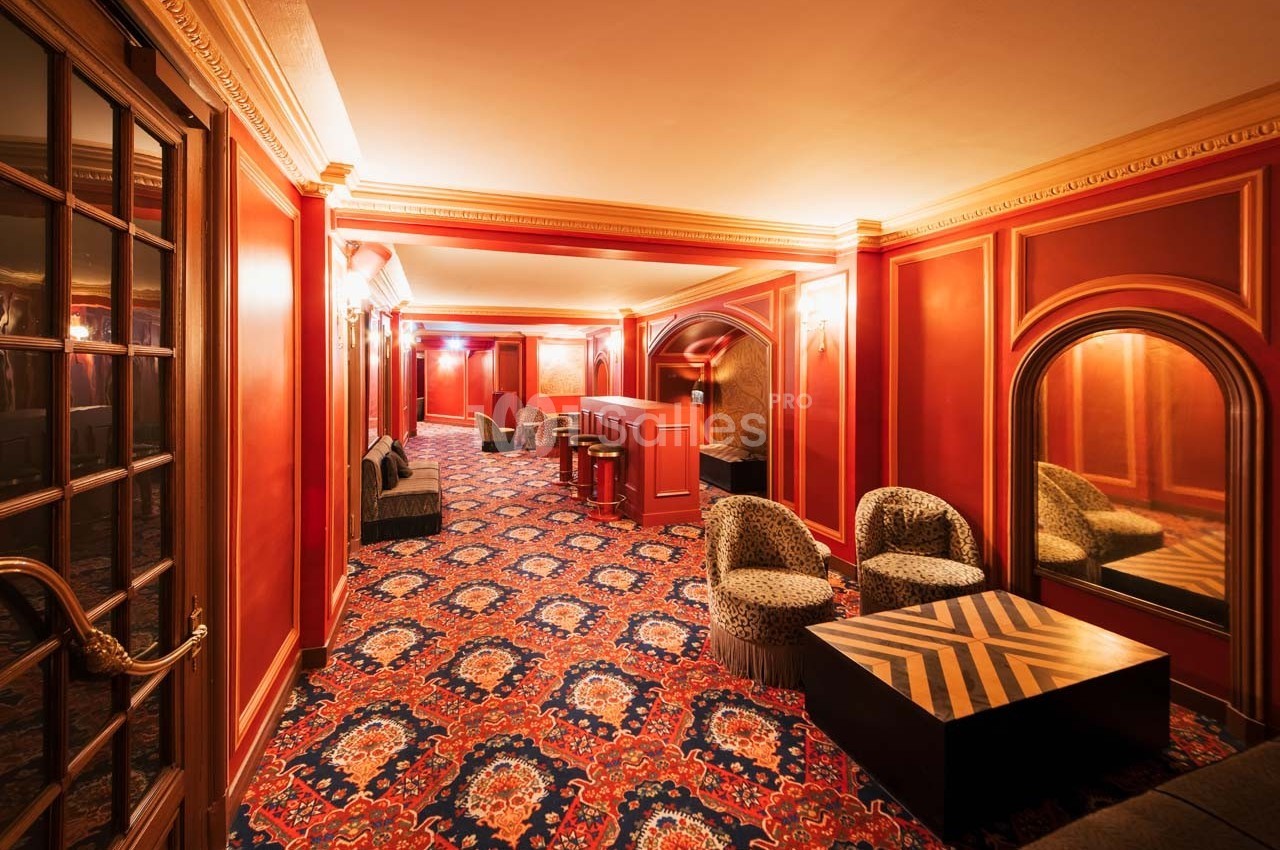 Salon élégant avec murs rouges, tapis à motifs, fauteuils, tables basses et miroirs dans un cadre chaleureux.